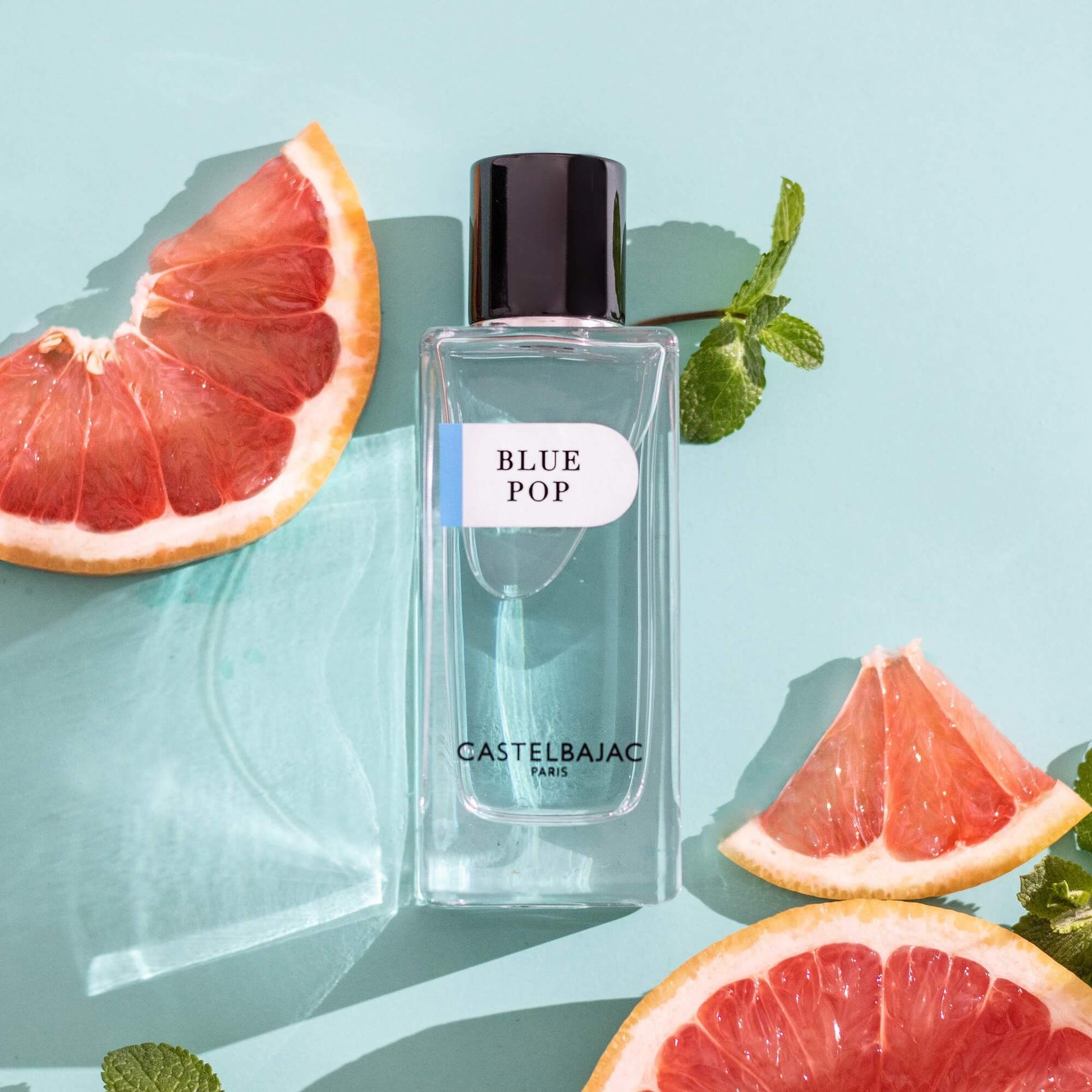 Castelbajac BLUE POP Eau de Parfum bottle with vibrant blue liquid, set against a backdrop of Mediterranean cliffs and turquoise waves